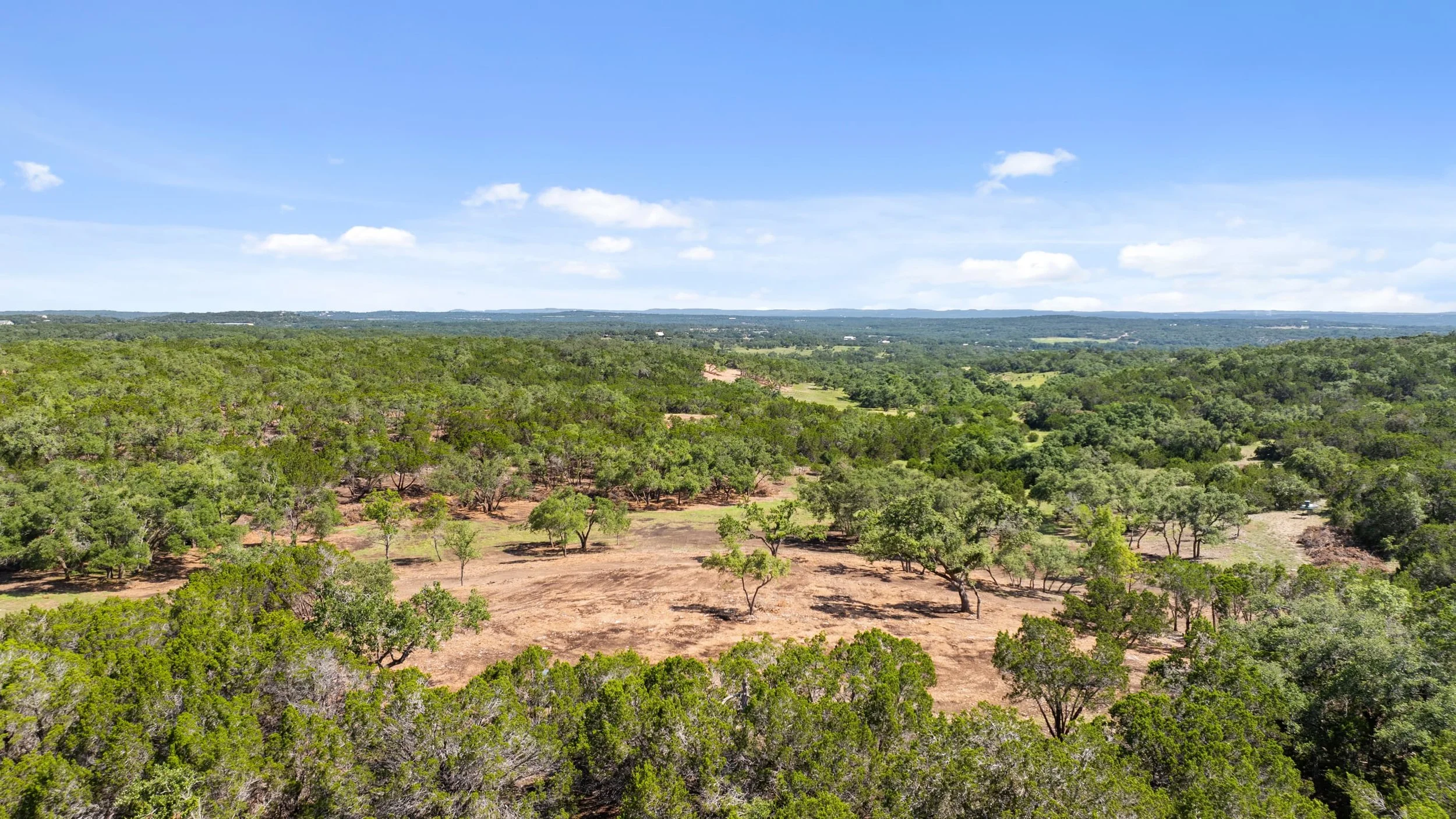 Aerial view of a green, semi-arid landscape with scattered trees and patches of bare soil, under a blue sky with some clouds.