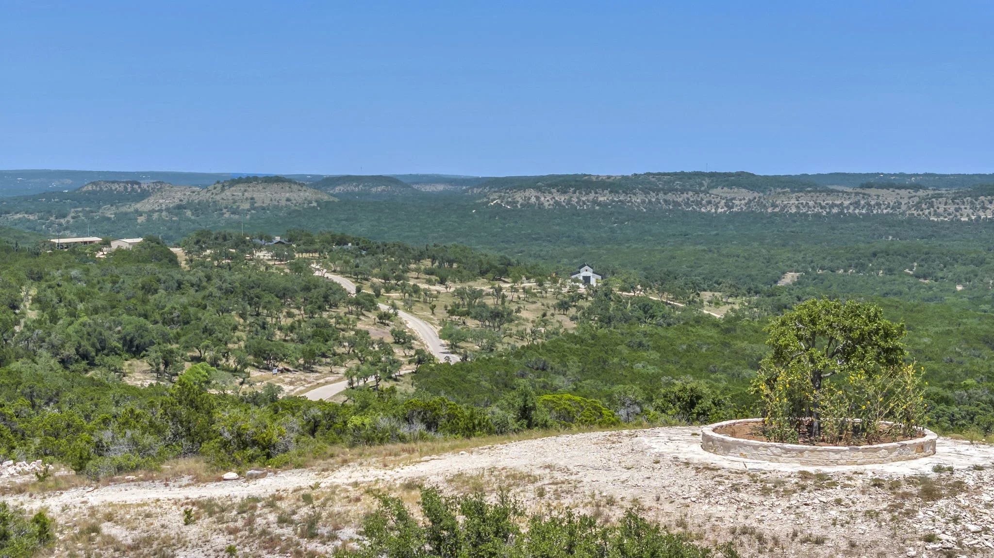 A rural landscape with a small tree in a circular raised brick planting bed in the foreground, rolling hills and green vegetation in the background, and a clear blue sky.