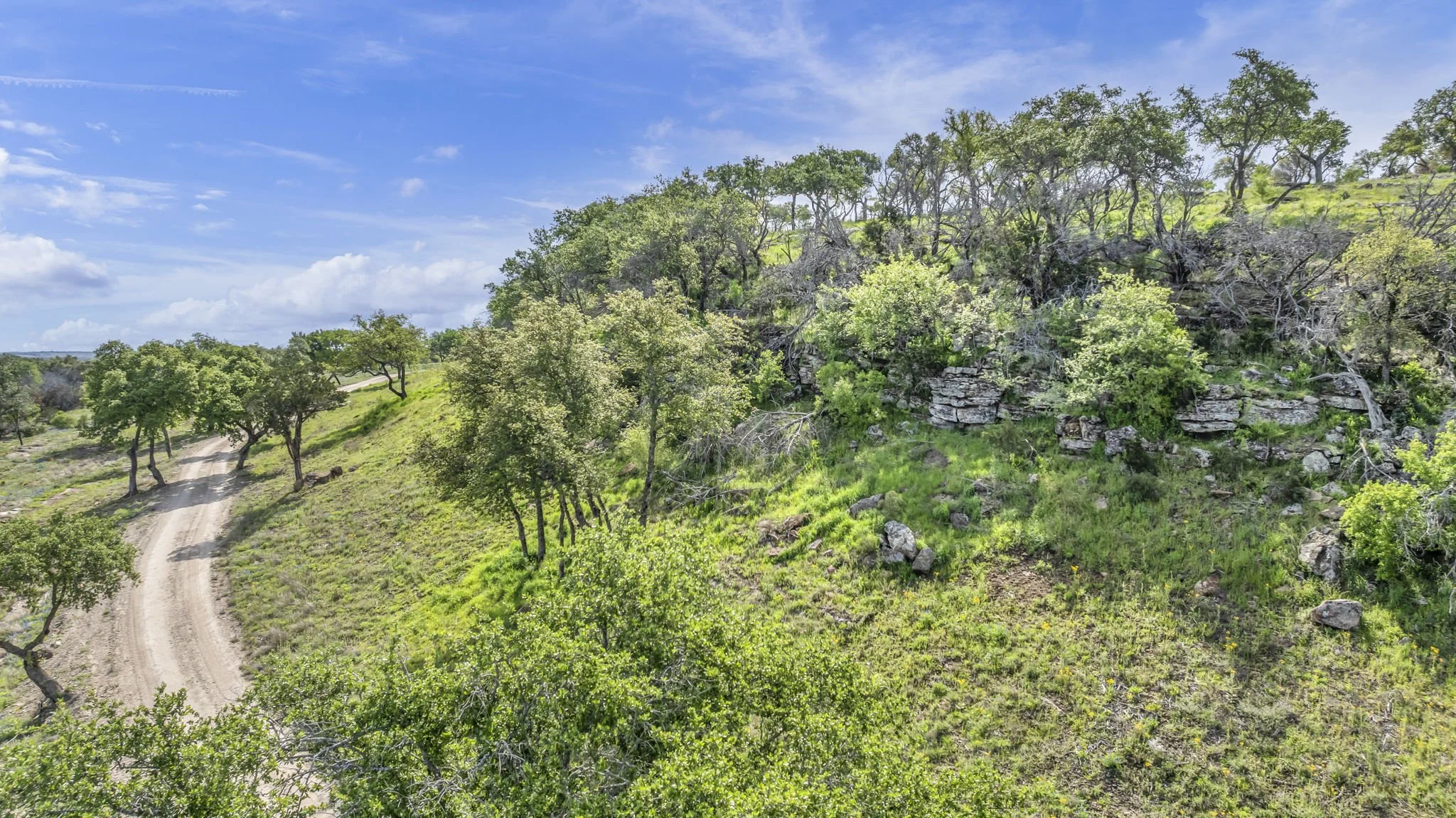 A dirt road winding through a green landscape with trees and rocky hills under a blue sky with scattered clouds.