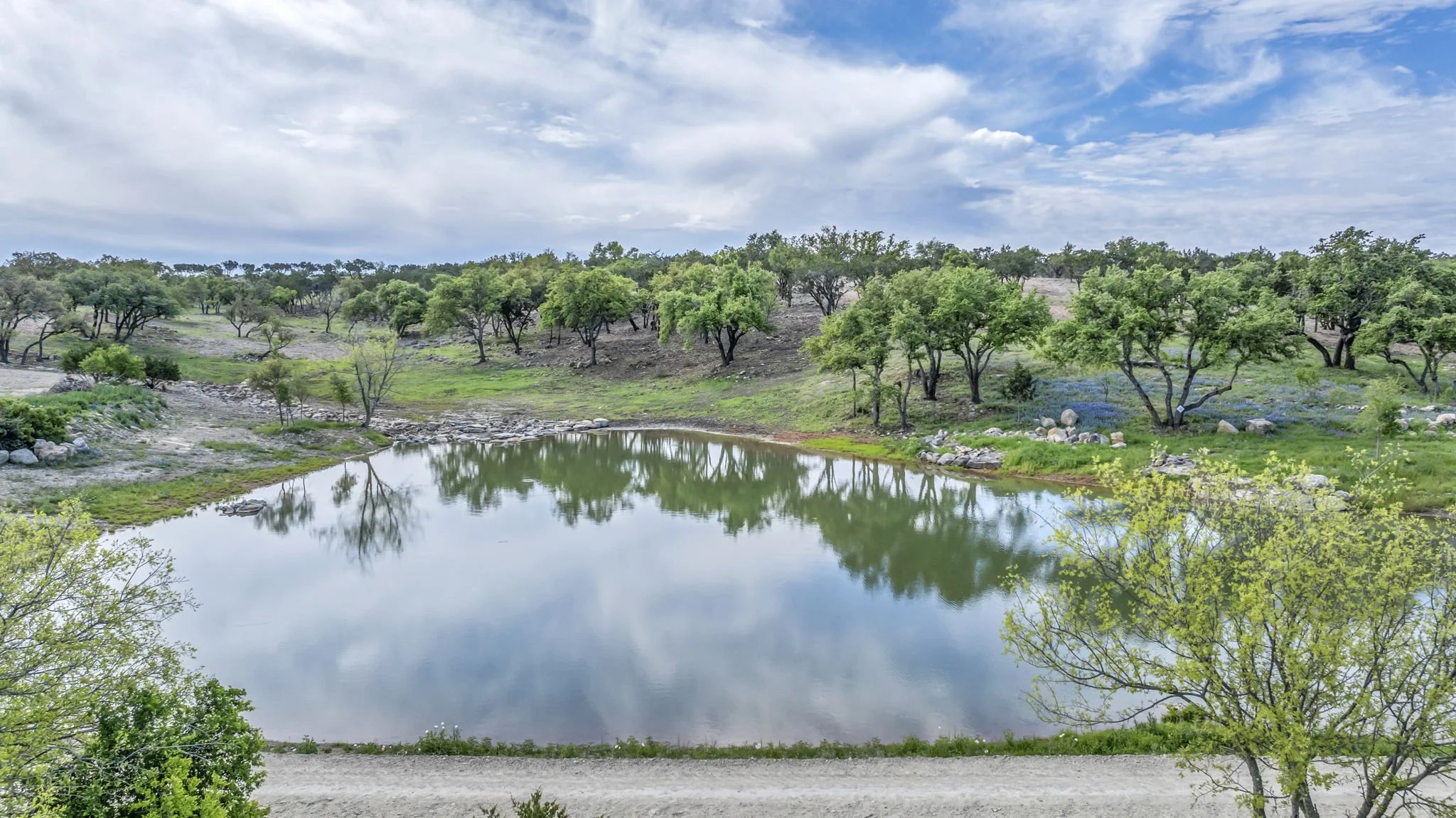 A serene landscape of a small lake surrounded by green trees and rolling hills, with a partly cloudy sky reflected in the water.